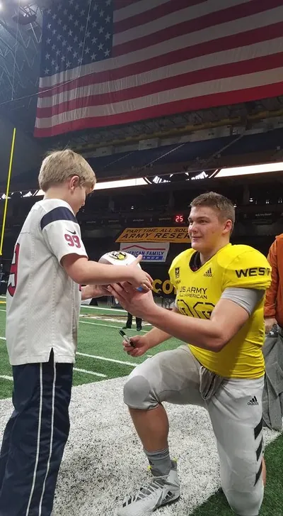 Draco Bynum in a yellow U.S. Army All-American jersey kneeling on the field to sign a football for a young fan, with a large American flag hanging in the stadium