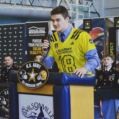 Draco Bynum in a yellow U.S. Army All-American No. 18 jersey over a shirt and tie, speaking at a U.S. Army All-American Bowl podium in the Wilsonville gym