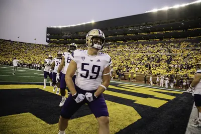 Draco Bynum in a white Washington No. 59 jersey and gold helmet, flexing in the end zone during warmups before Washington's game at Michigan Stadium on Sept. 11, 2021, with a packed crowd in maize behind him