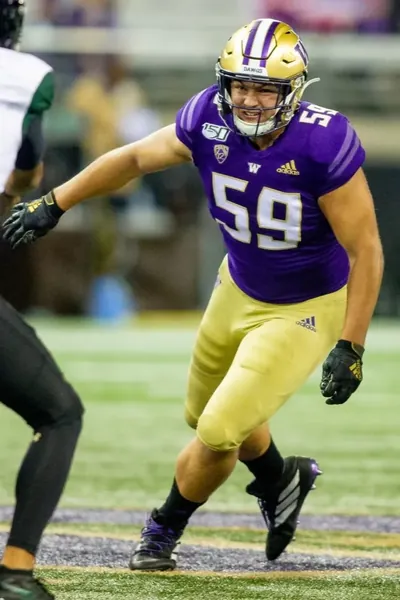 Draco Bynum in a purple Washington No. 59 home jersey, gold pants and gold helmet, pass rushing against a Hawaii player in a white jersey during Washington's home game against Hawaii at Husky Stadium on Sept. 14, 2019