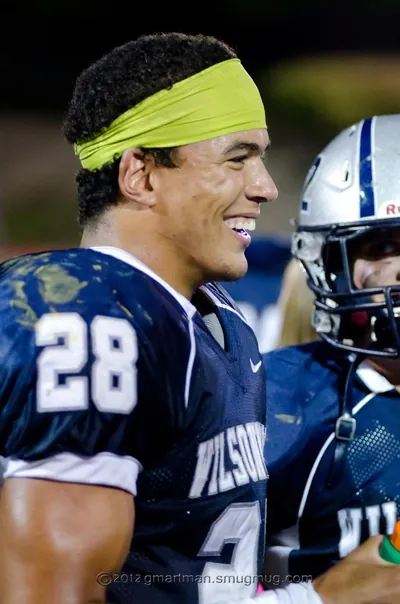 Johnny Ragin III, Wilsonville No. 28, smiling on the sideline at a night game in a navy jersey and yellow headband, 2012