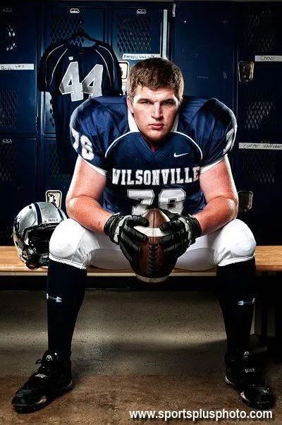 Andrew Walls in his navy Wilsonville No. 76 uniform, seated on a locker-room bench holding a football, with his helmet beside him